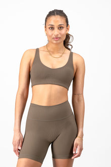 A woman modeling a matching athletic set consisting of a cropped sports bra and high-waisted biker shorts in a solid taupe-brown color, shot against a clean white background.
